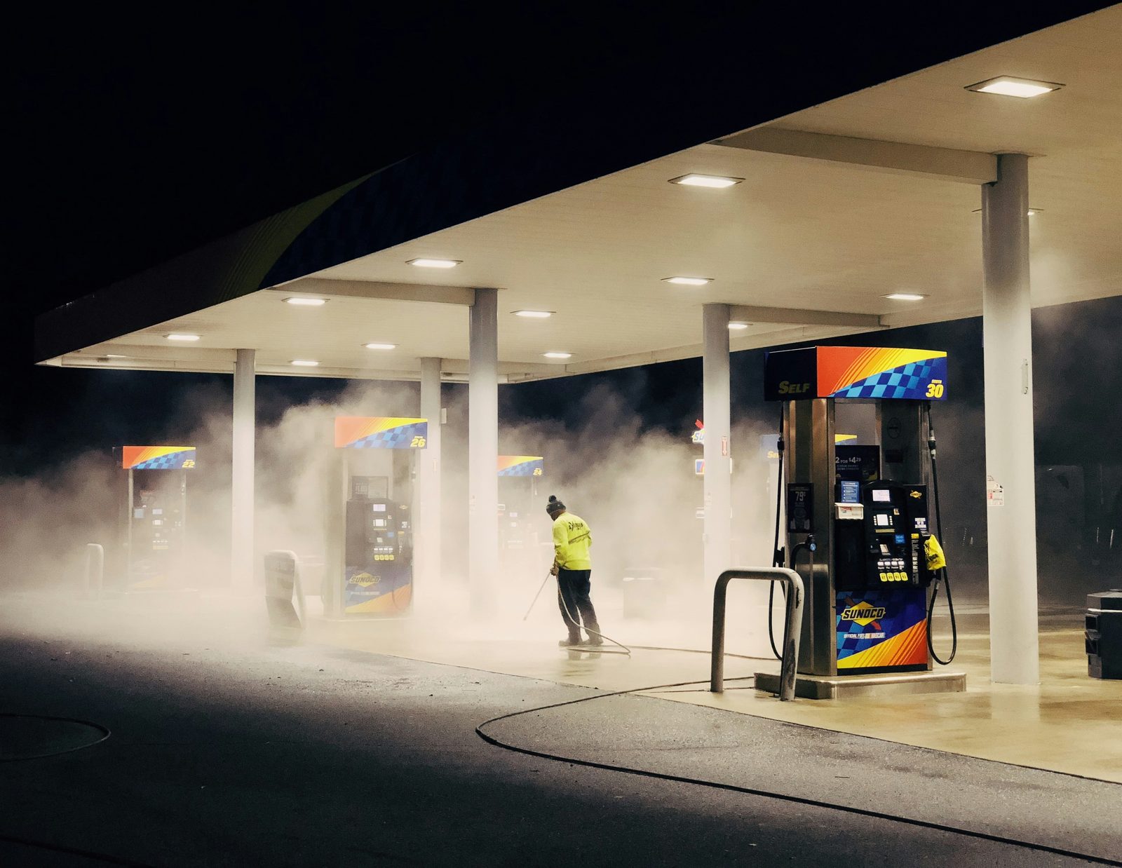 Pressure washing a commercial gas station forecourt at night, steam rising off the concrete